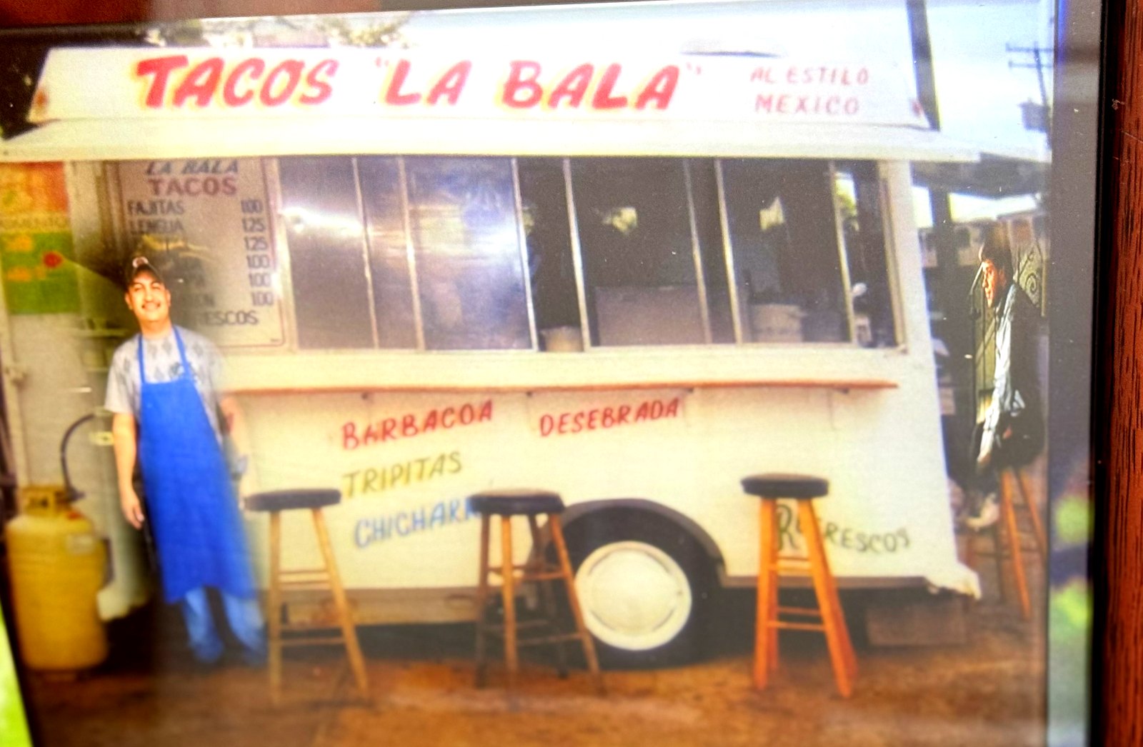 The original Tacos La Bala trailer with the name hand-painted across the top, the founder standing beside it in a blue apron